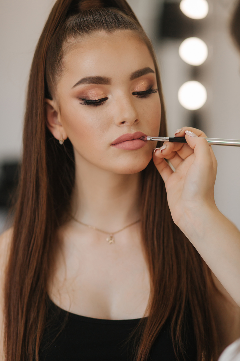 Makeup artist applies lipstick. Hand of make-up master, painting lips of young beauty redhead model. Make up in process